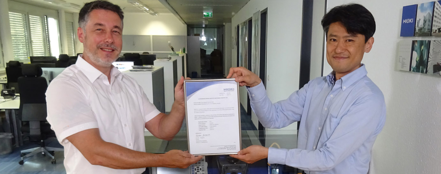 Zwei Mitarbeiter halten die Urkunde der Zusammenarbeit lächelnd in die Kamera.